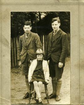 Two boys standing and young girl sat on chair